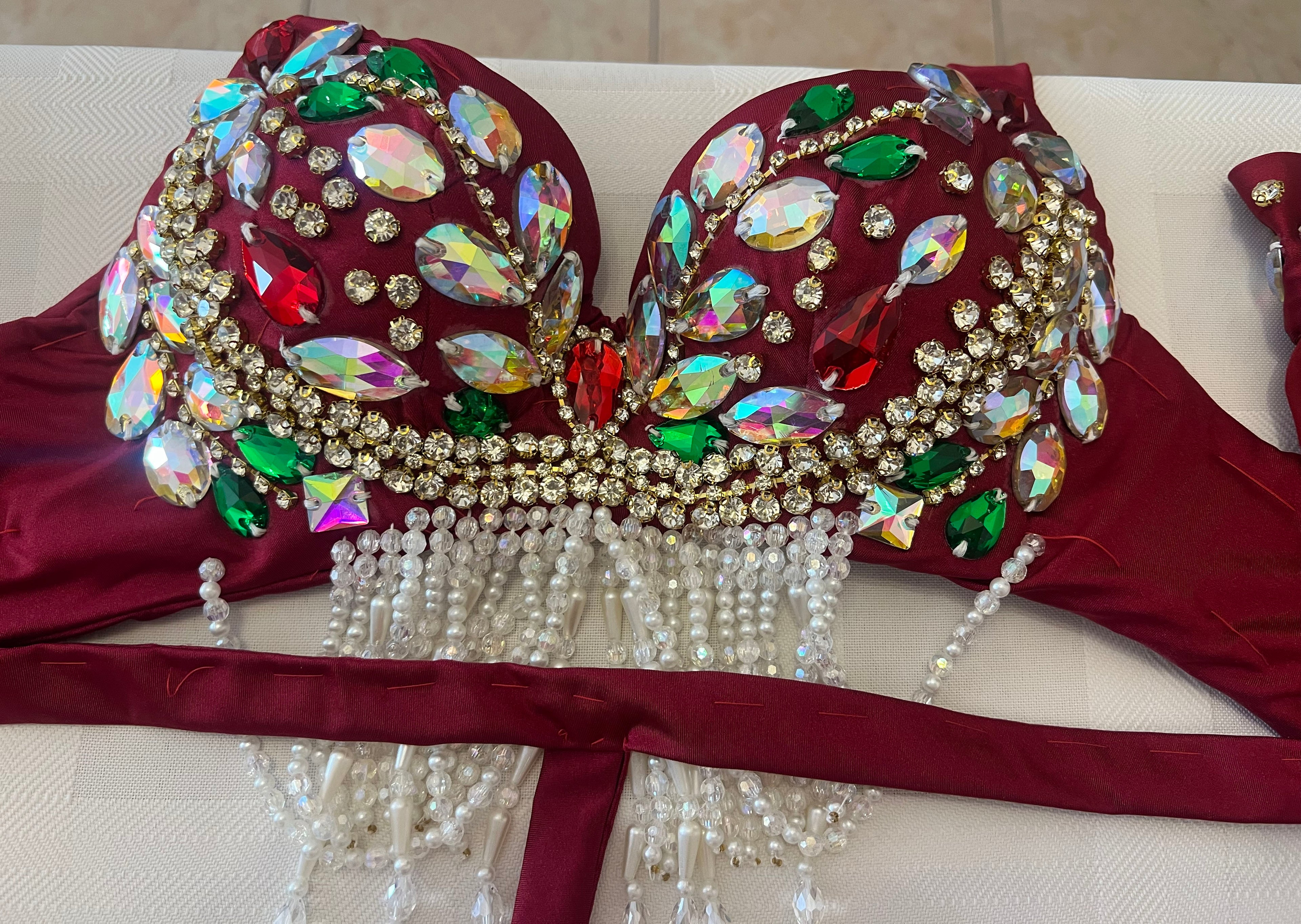 A close-up of a burgundy belly dance bra adorned with large iridescent rhinestones, green and red crystals, gold rhinestone borders, and dangling white pearl bead strands, laid flat on a white surface.