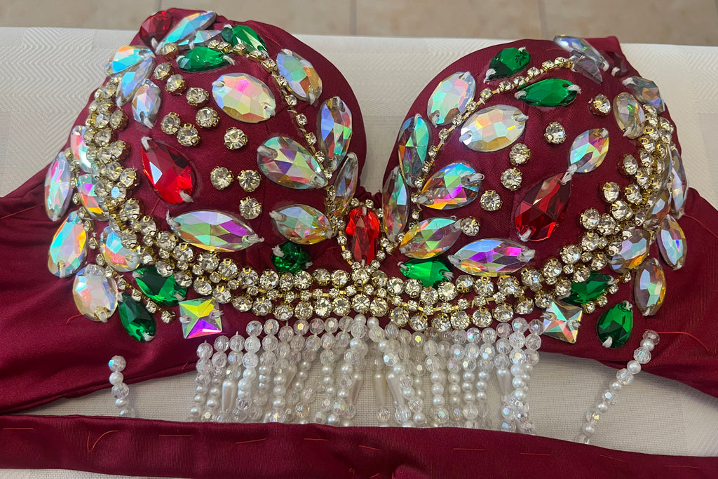 A close-up of a heavily embellished dance or costume bra top in deep burgundy, decorated with large iridescent rhinestones, green and red gemstones, gold rhinestone borders, and dangling white pearl bead fringes.