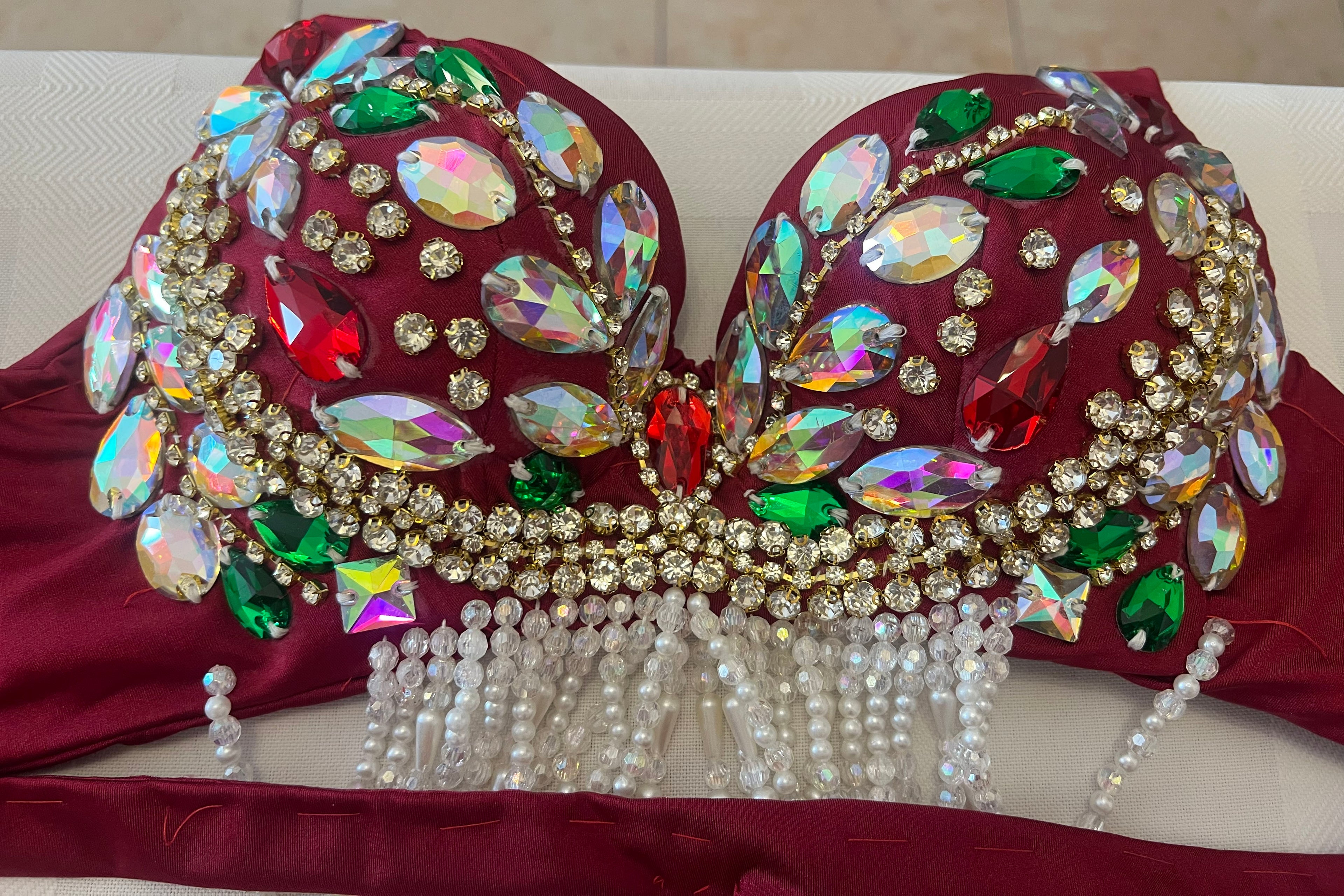 A close-up of a heavily embellished dance or costume bra top in deep burgundy, decorated with large iridescent rhinestones, green and red gemstones, gold rhinestone borders, and dangling white pearl bead fringes.