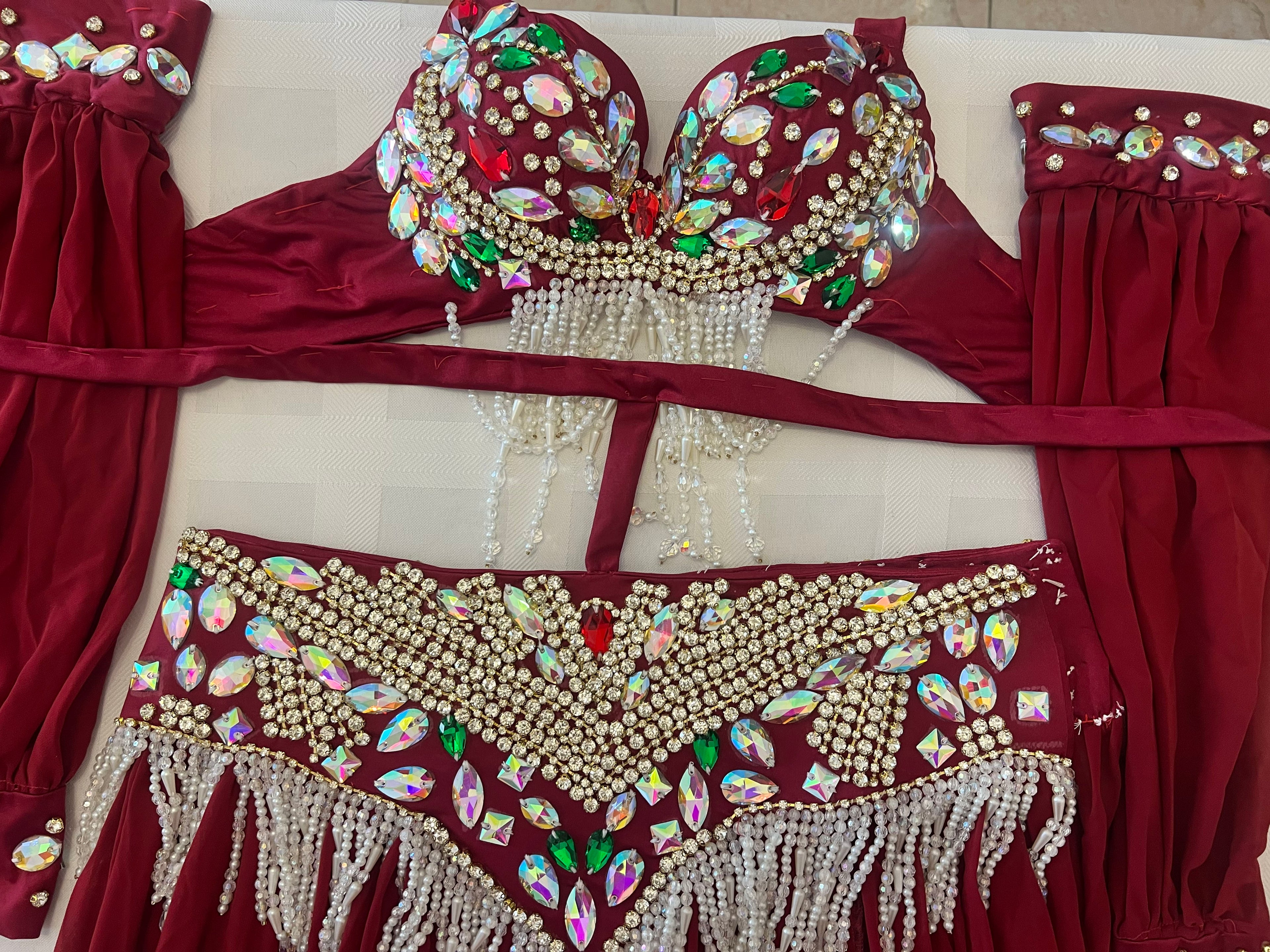 A two-piece belly dance costume set in deep burgundy velvet, featuring a strapless bra top and matching bottoms, both heavily embellished with iridescent rhinestones, white faux pearls, and dangling bead chains, laid flat on a light-colored surface.