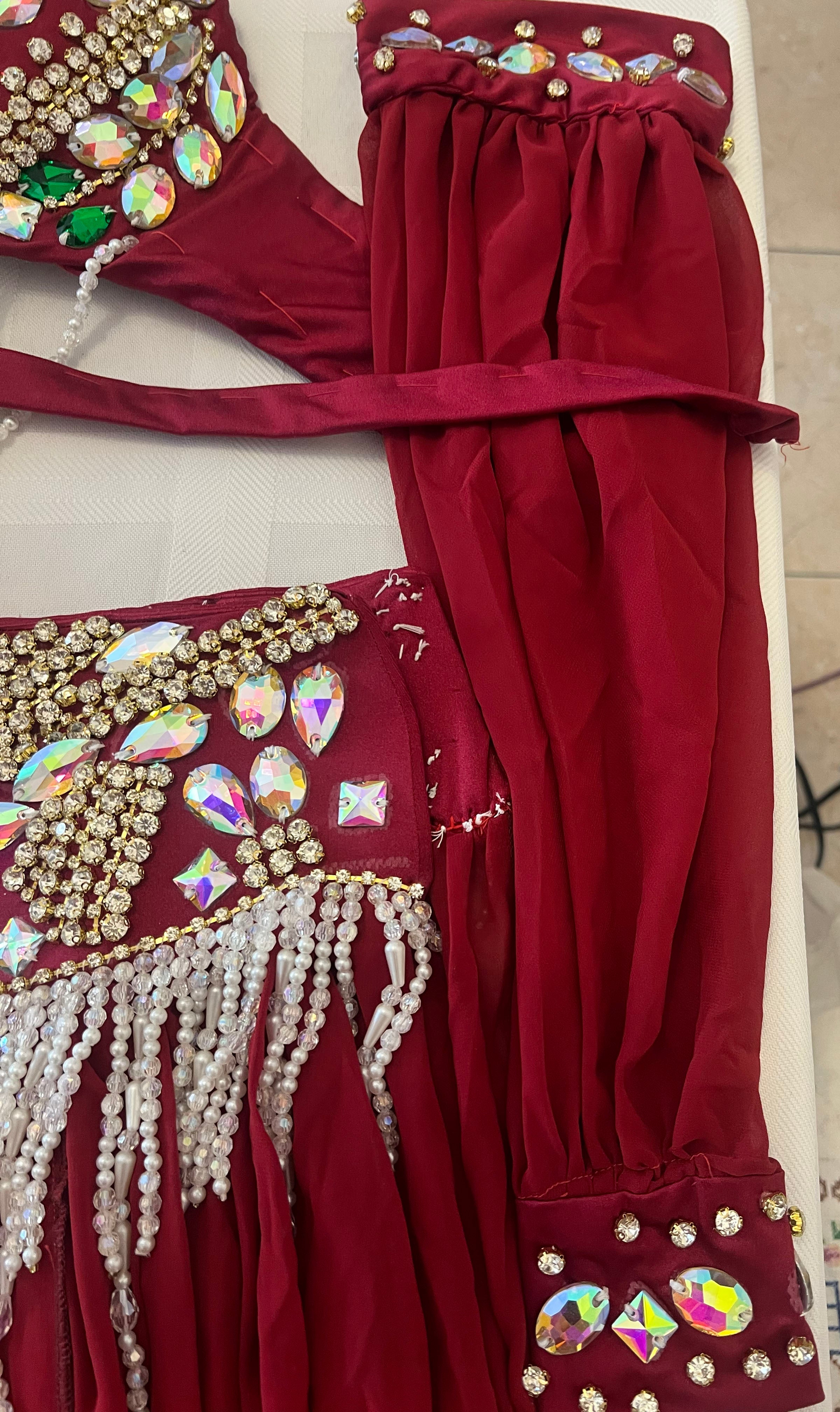 A close-up of a deep red belly dance costume featuring intricate gold and iridescent rhinestone embellishments, white bead fringe, and matching arm warmers on a light-colored surface.