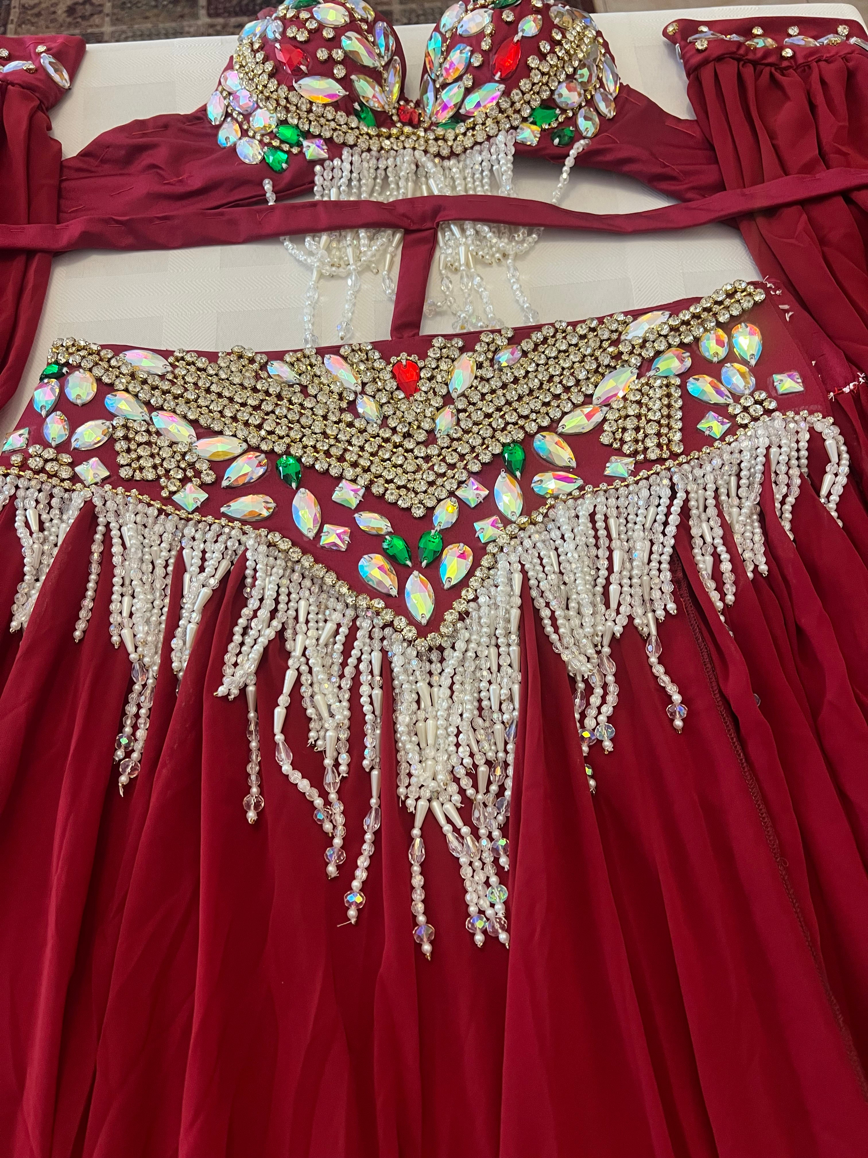 A red belly dance costume set featuring a heavily embellished bra top and matching hip scarf, both decorated with gold coins, colorful rhinestones, and long white bead fringes, laid flat on a light surface.