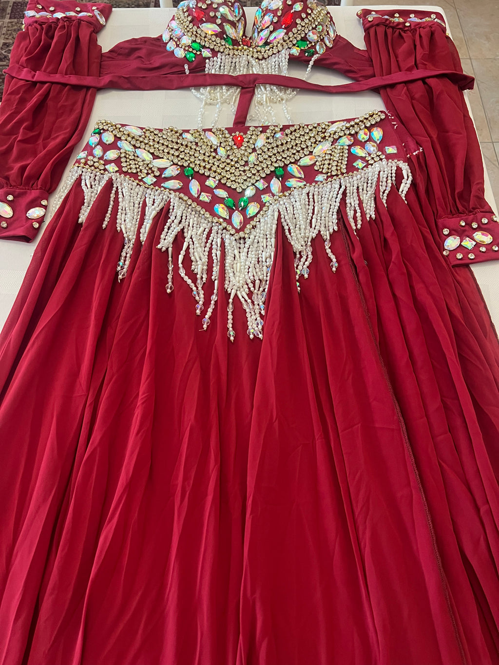 A red off-the-shoulder dress with extensive gold beadwork, white beaded fringe, and colorful embellishments, shown laid flat with a matching embroidered waistband.
