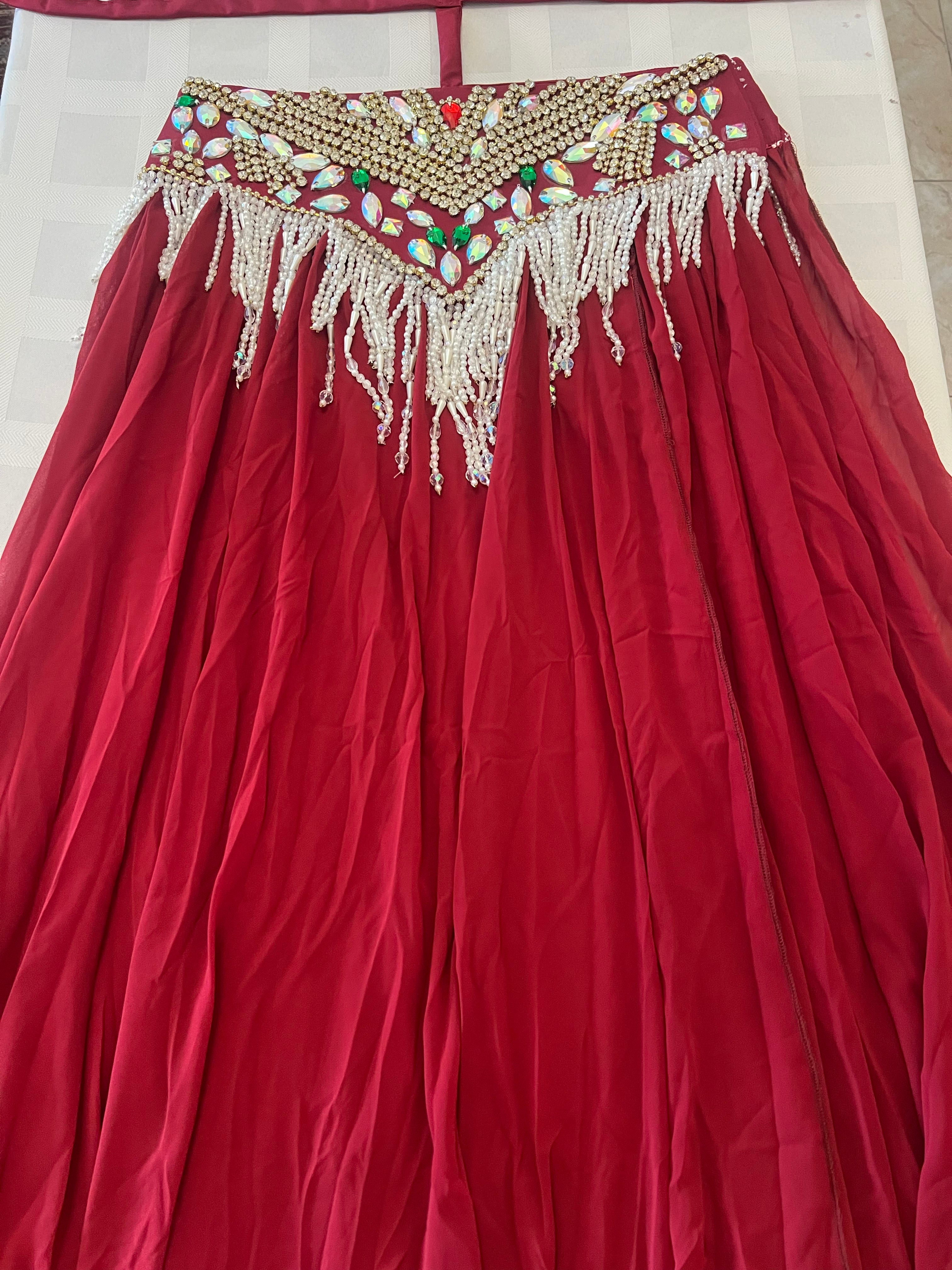 A red traditional skirt or lehenga with an ornate embroidered waistband, featuring gold beadwork, colorful mirror work, and white beaded fringe, laid flat on a light-colored surface.