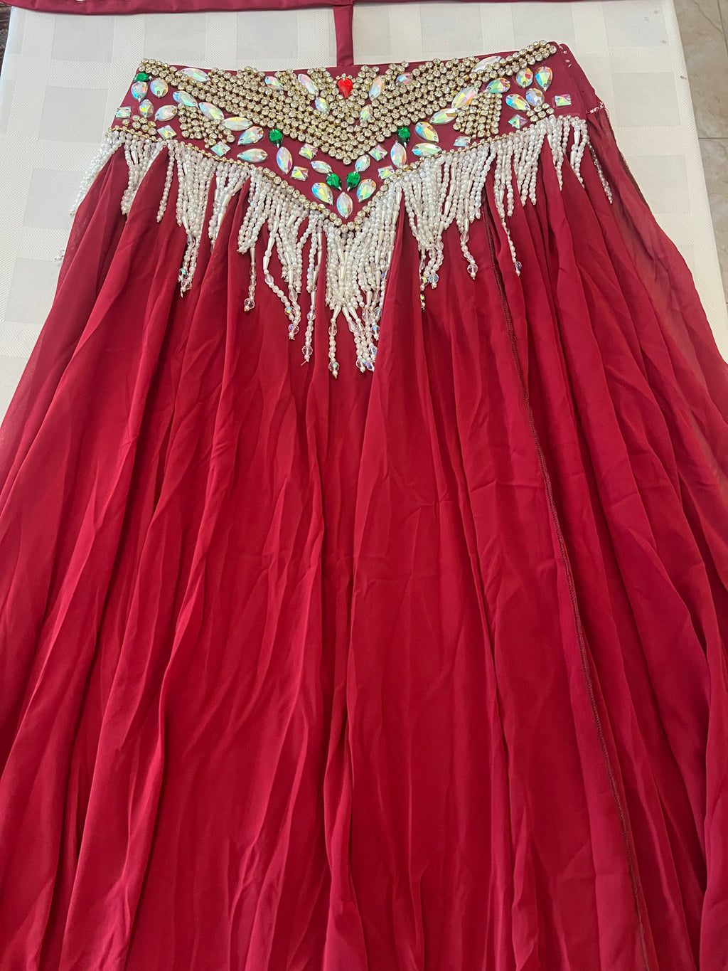 A red traditional skirt or lehenga with an ornate embroidered waistband featuring gold beadwork, multicolored stones, and white beaded fringe, laid flat on a light surface.