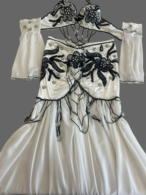 White belly dance bra and skirt set with black floral beadwork and matching chiffon sleeves, laid flat on gray