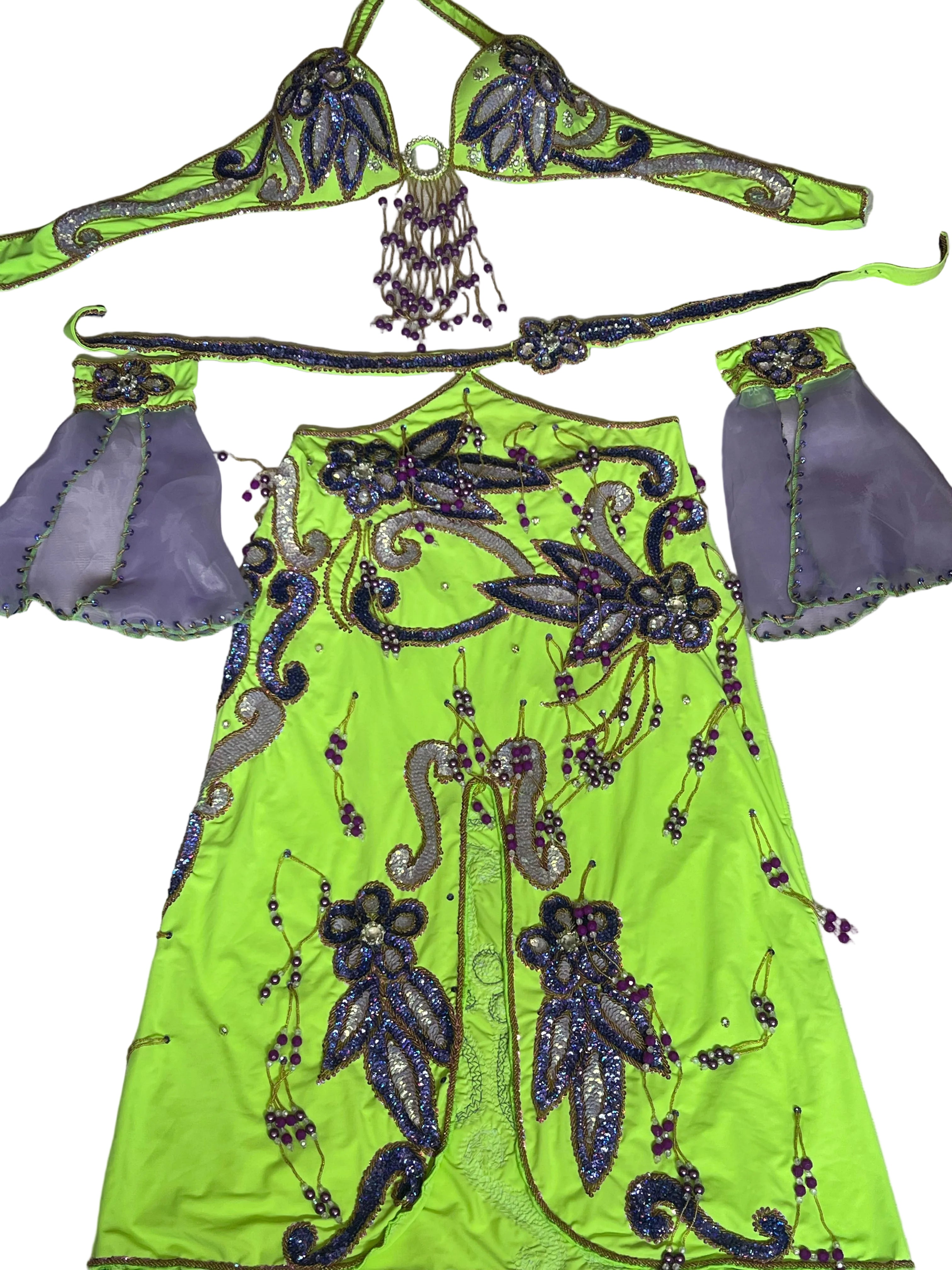 A neon yellow belly dance costume set laid flat on a white fabric background, featuring a beaded top with dangling beads, a matching belt, purple sheer sleeves, and a long skirt with purple and silver beadwork.