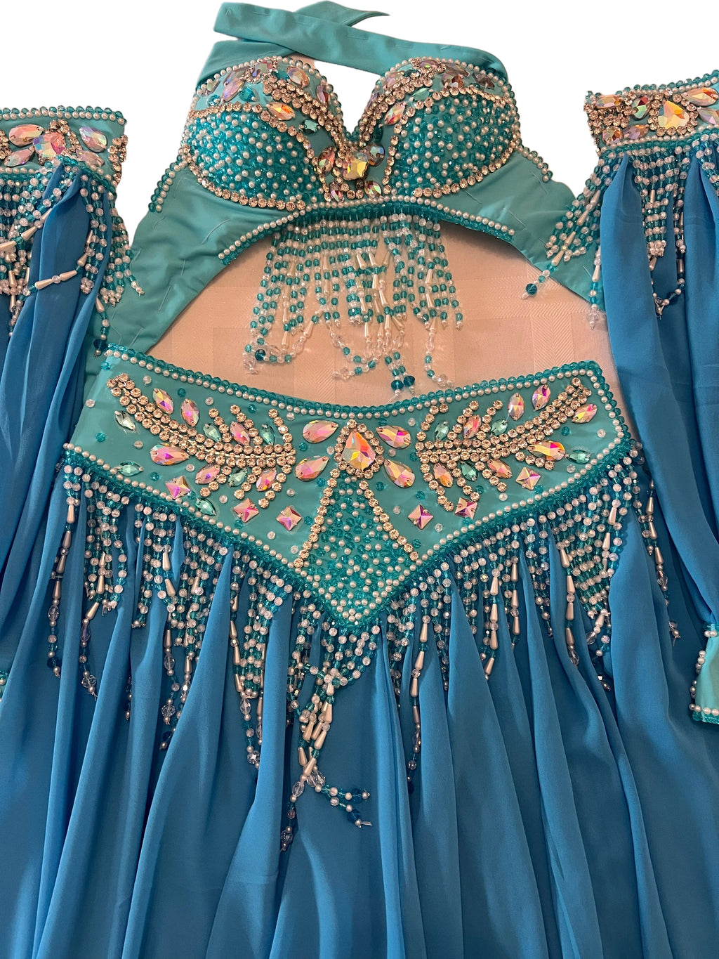 A turquoise and blue belly dance costume set with intricate gold beadwork, rhinestones, and dangling beaded fringe, laid out on a light wood floor.