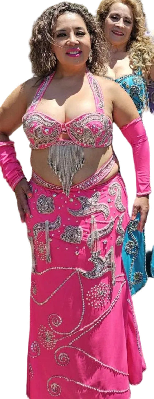 A woman wearing a hot pink Egyptian belly dance costume with embellishments including large clear glass stones, glass beads, and sequins. The costume includes a skirt, bra, sleeves, and a headband.