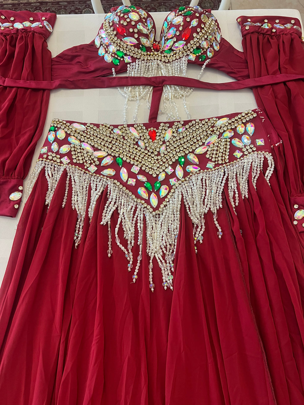 A red belly dance costume set featuring a long pleated skirt with heavy gold beadwork and white fringe, a matching bra top with colorful jewel embellishments, and coordinating sleeve cuffs, laid out on a light surface.