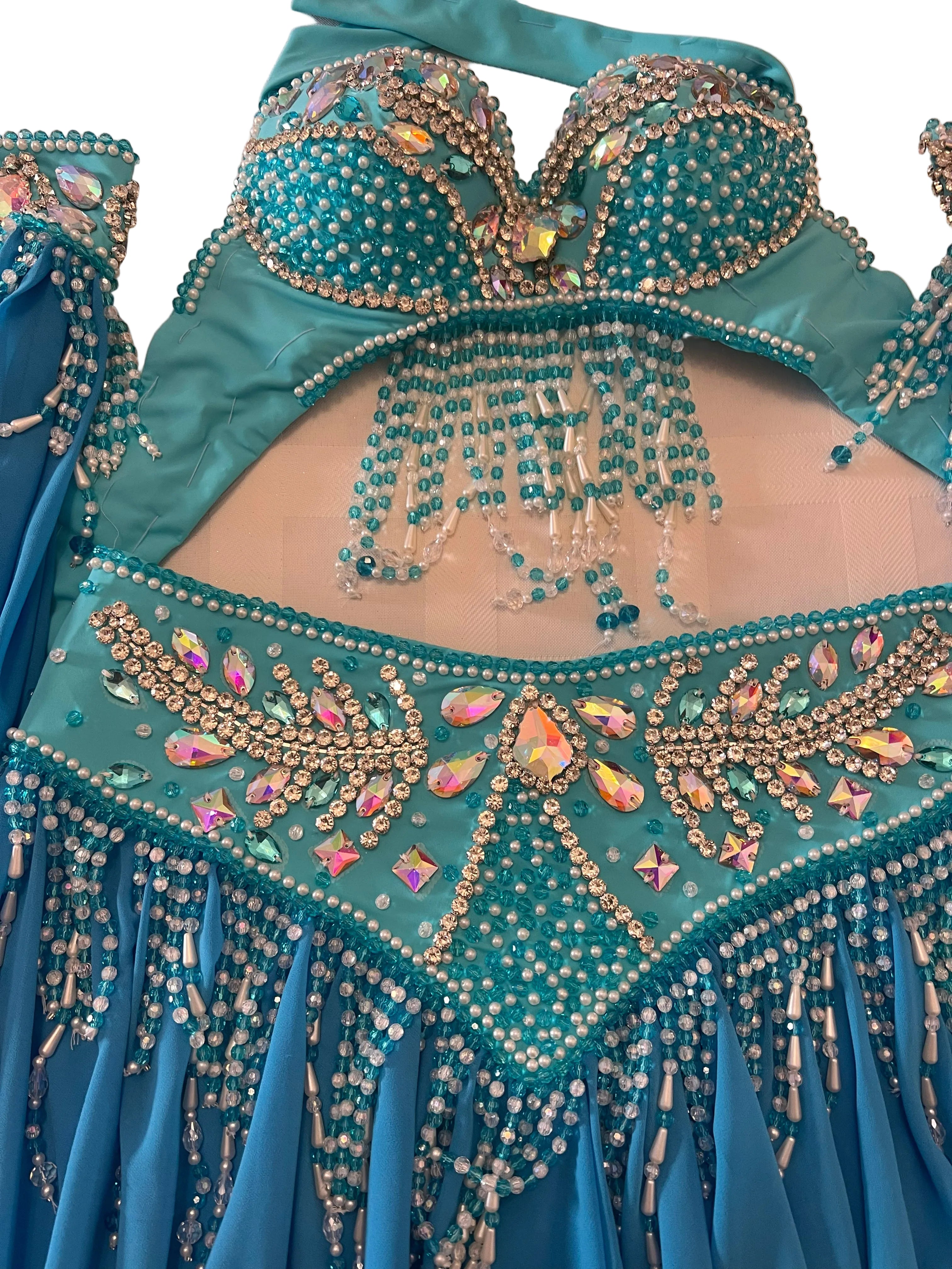 A turquoise belly dance costume set featuring a heavily embellished bra top with dangling rhinestone fringe, a matching hip scarf with large decorative crystals, and a blue pleated skirt with bead and crystal fringe.