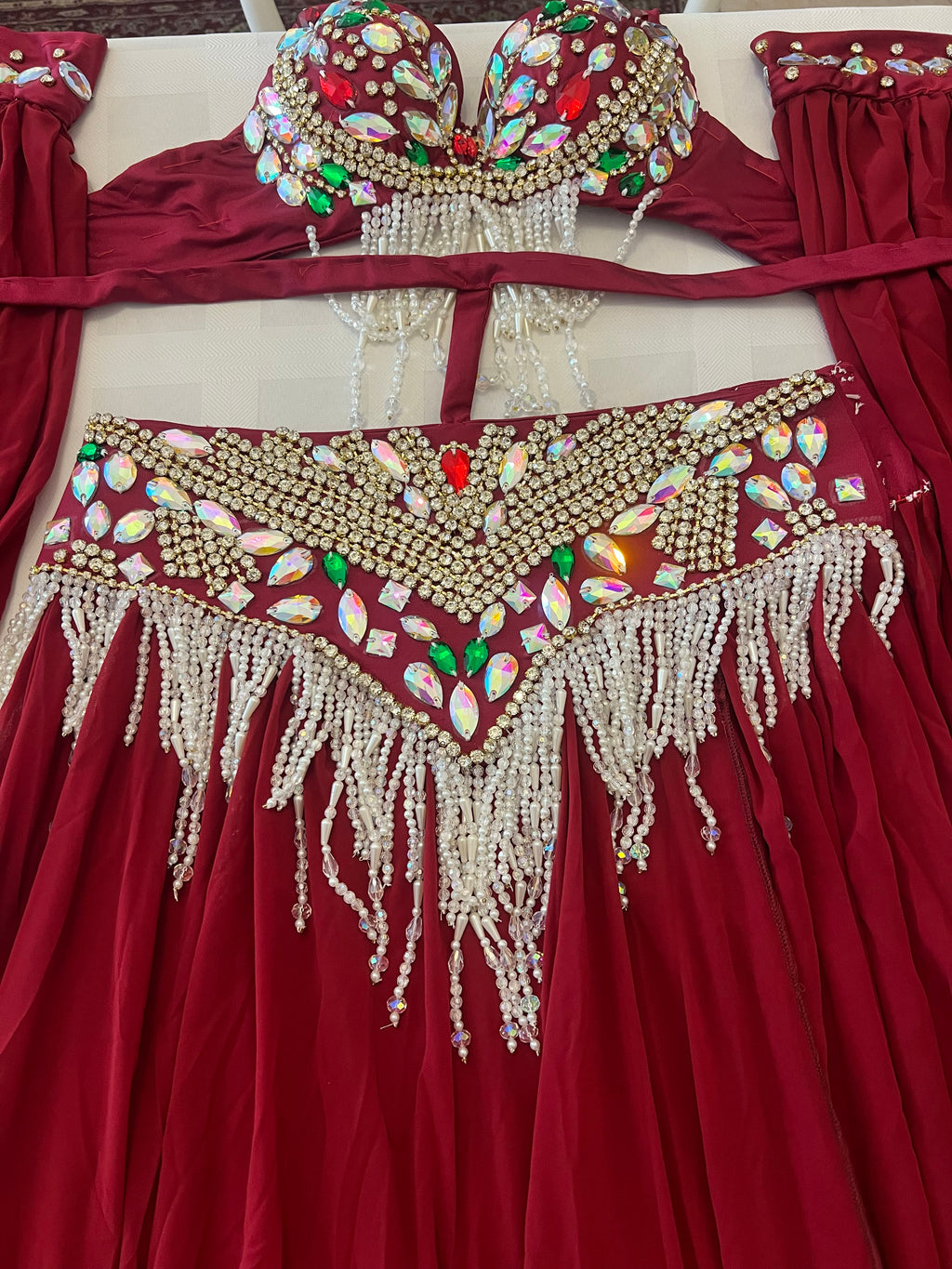 A red belly dance costume set featuring a heavily embellished bra top and matching hip scarf, both decorated with gold beads, white pearl-like fringe, and colorful rhinestones, laid flat on a light surface.