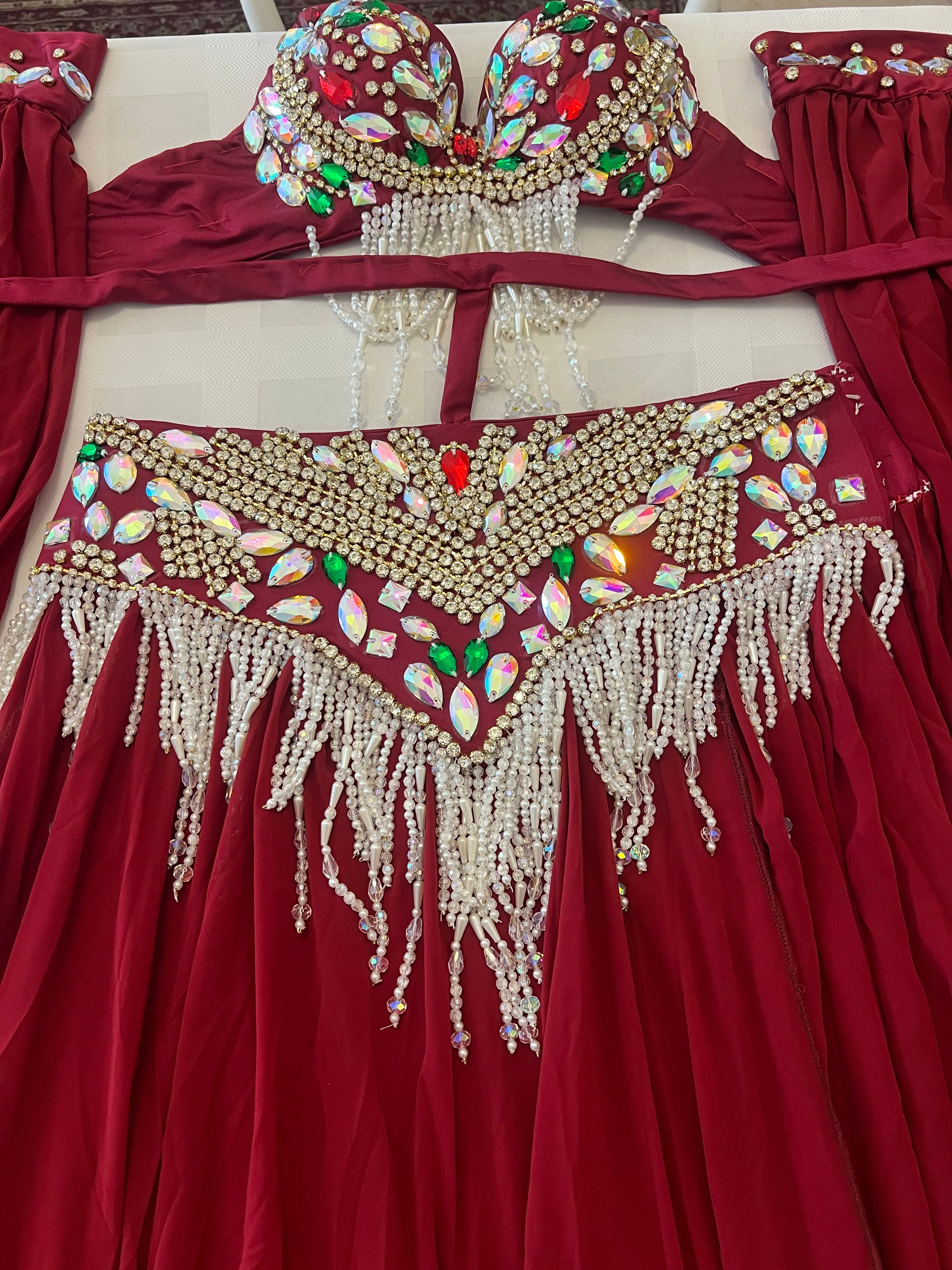 A red belly dance costume set featuring a heavily embellished bra top and matching hip scarf, both decorated with gold beads, white pearl-like fringe, and colorful rhinestones, laid flat on a light surface.