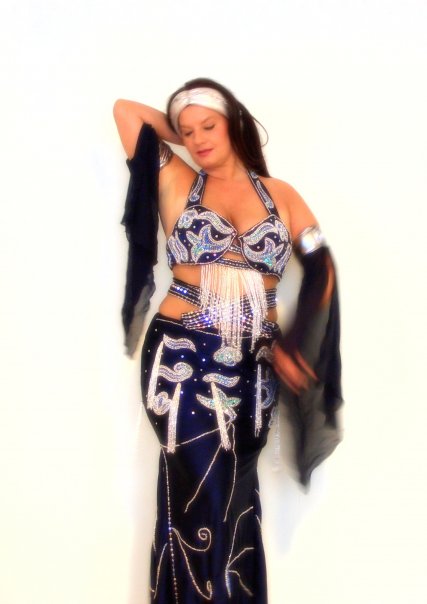 A woman modeling a custom-made belly dance costume with a black skirt, blue accents, and geometric patterns. The costume includes a headband and the model is posing with her hand behind her head.