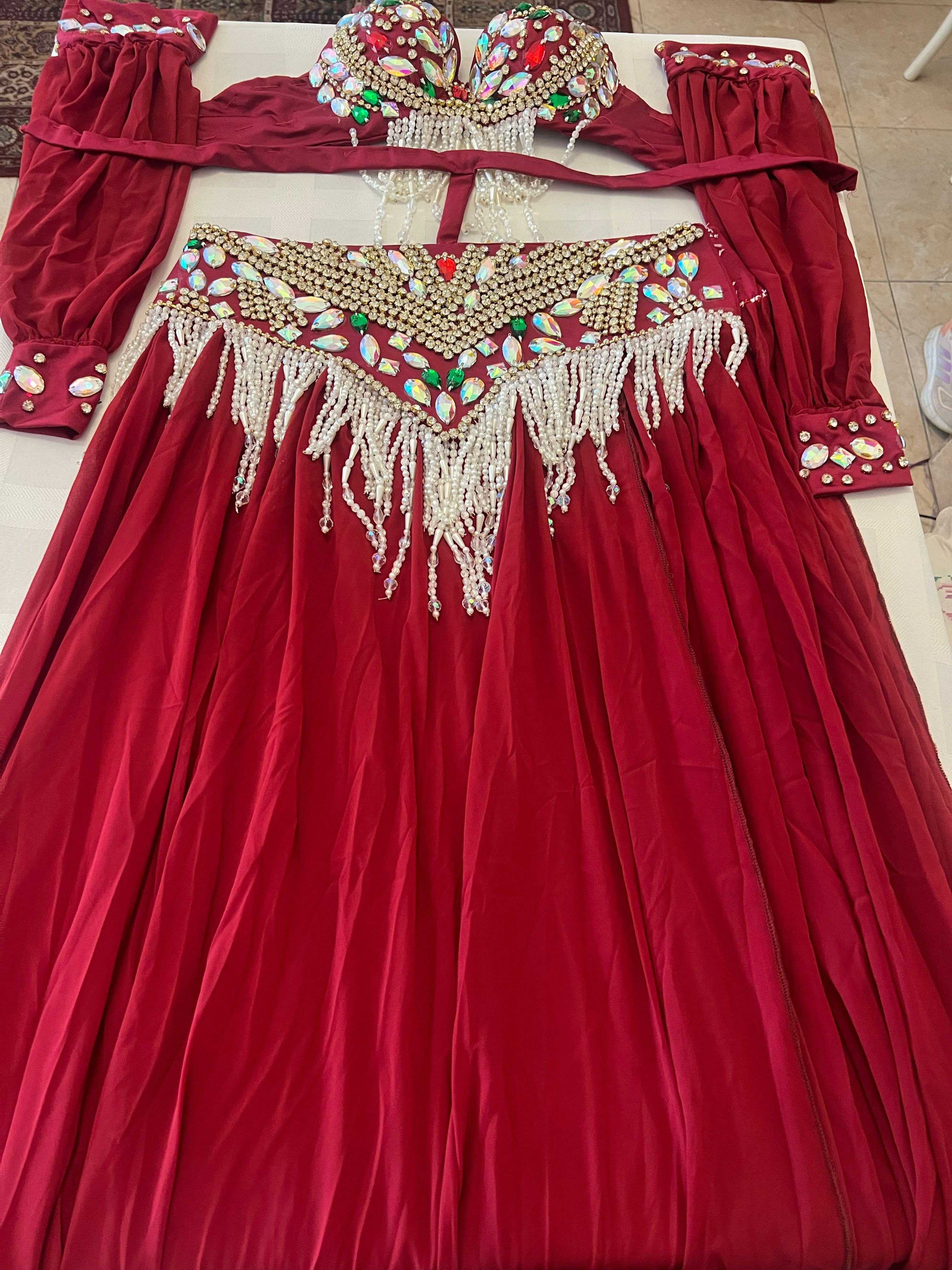 A red belly dance costume laid flat on a light-colored floor, featuring a long skirt with a beaded and fringed neckline, matching long sleeves with decorative cuffs, and a matching headpiece.