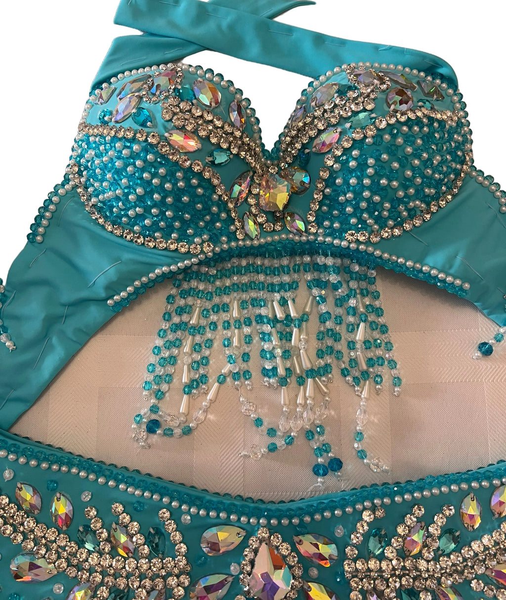 Turquoise belly dance costume set featuring a bra top and matching bottoms, heavily embellished with rhinestones, beads, and iridescent crystals on a white textured surface.