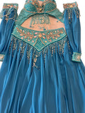 A turquoise blue belly dance costume laid flat on a light beige surface, featuring a long-sleeved skirt, a beaded bra top, and a matching hip scarf with intricate gold, silver, and pink rhinestone embellishments.