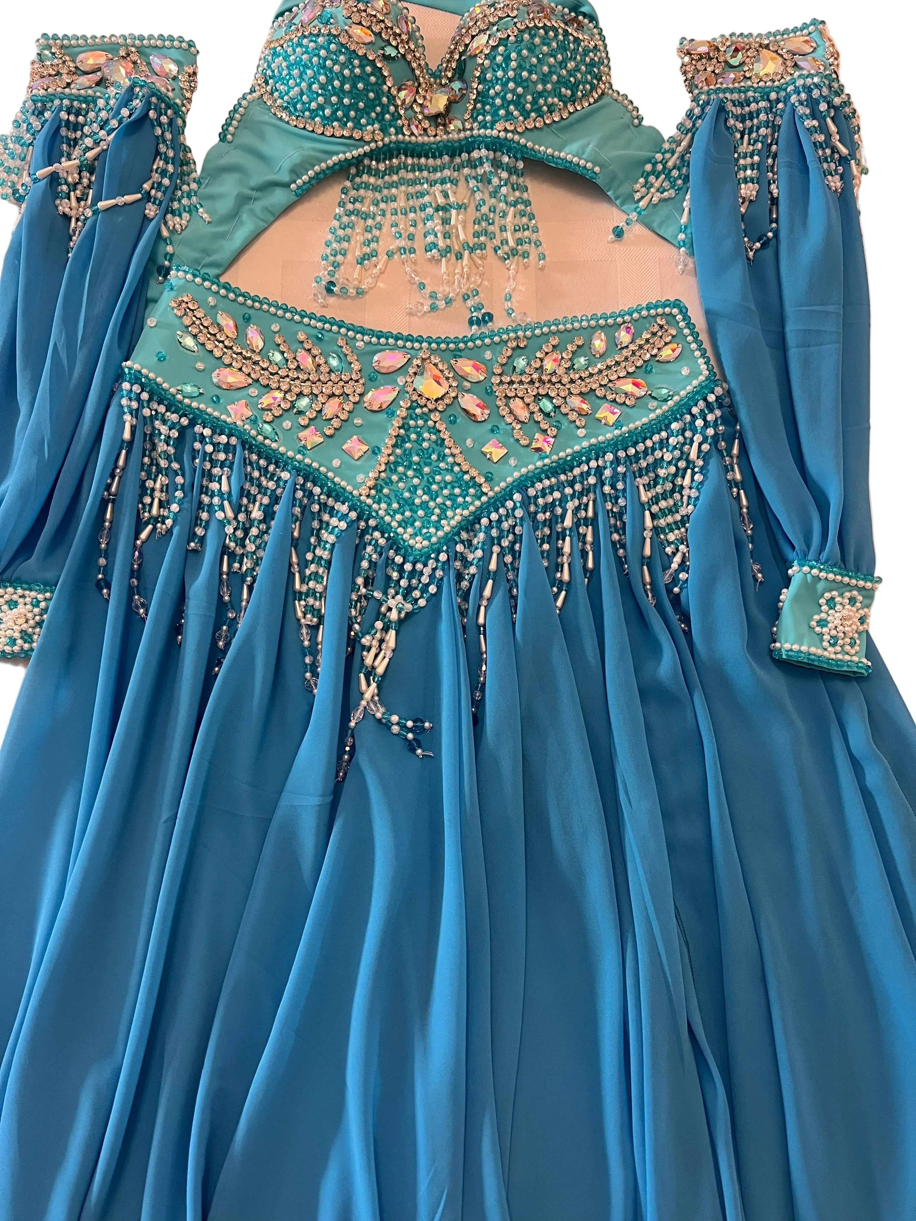 A turquoise blue belly dance costume laid flat on a light beige surface, featuring a long-sleeved skirt, a beaded bra top, and a matching hip scarf with intricate gold, silver, and pink rhinestone embellishments.