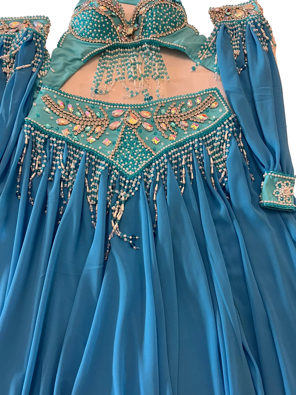 A turquoise blue belly dance costume set with intricate gold, silver, and white beadwork and sequins, laid flat on a light beige surface.