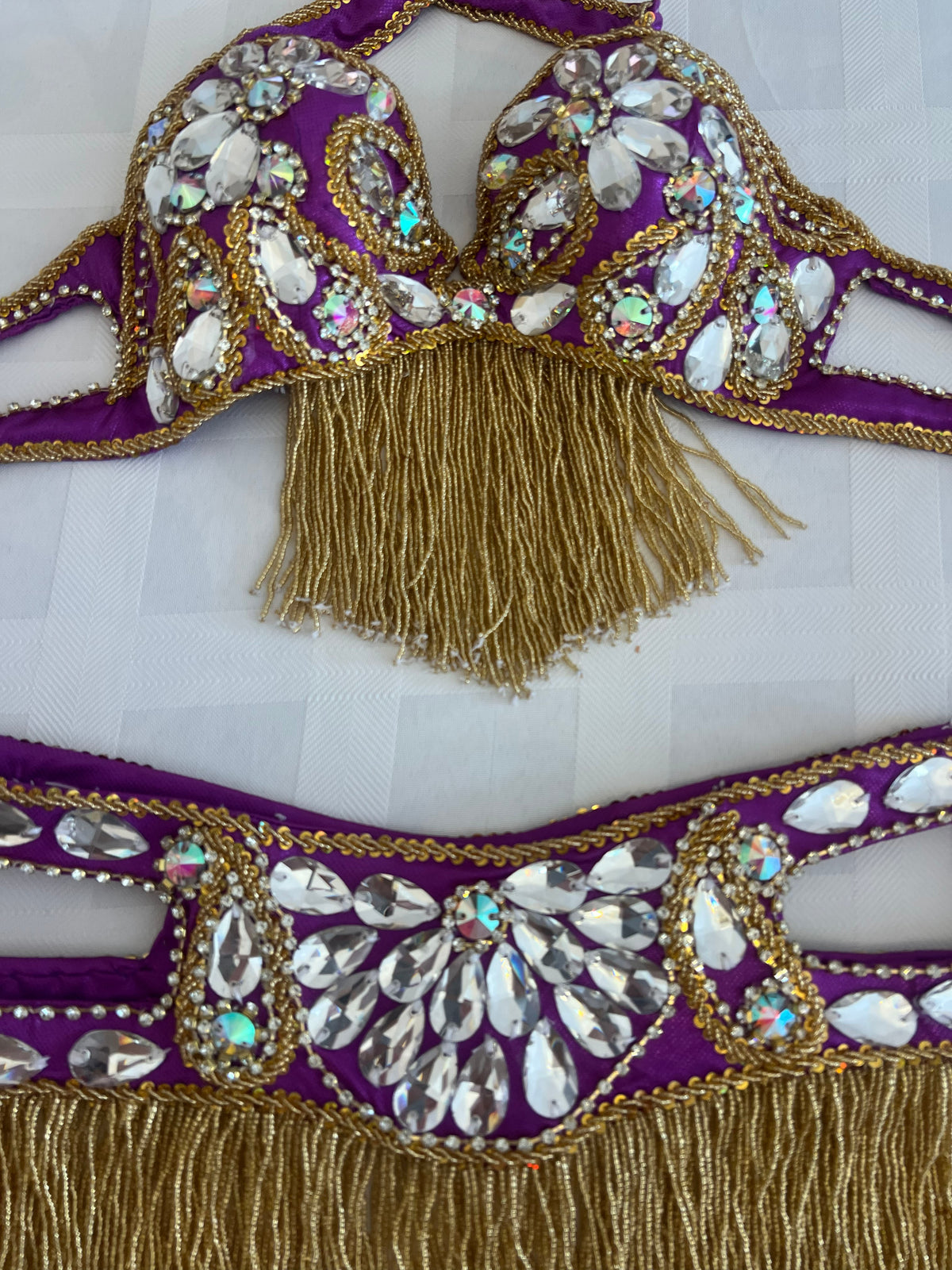 Purple and gold embellished dance costume set featuring a bra top with fringe and a matching hip scarf, both decorated with rhinestones and sequins on a light gray background.