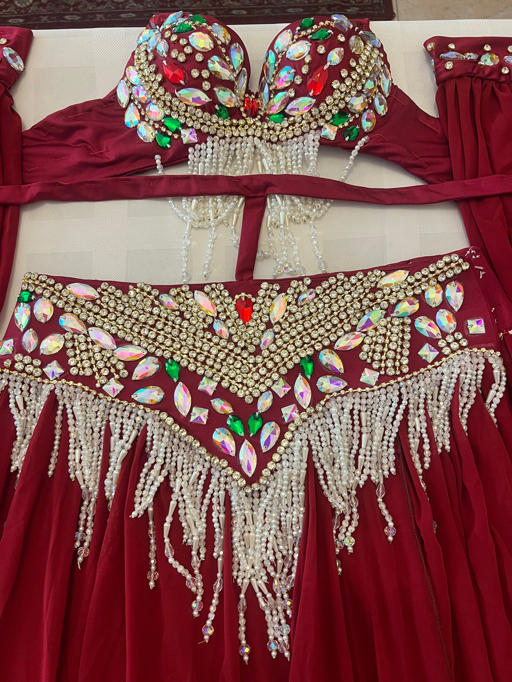 A close-up of a red belly dance costume set featuring a bra top and hip scarf with extensive gold rhinestone embellishments, iridescent teardrop gems, and dangling white pearl bead fringes.