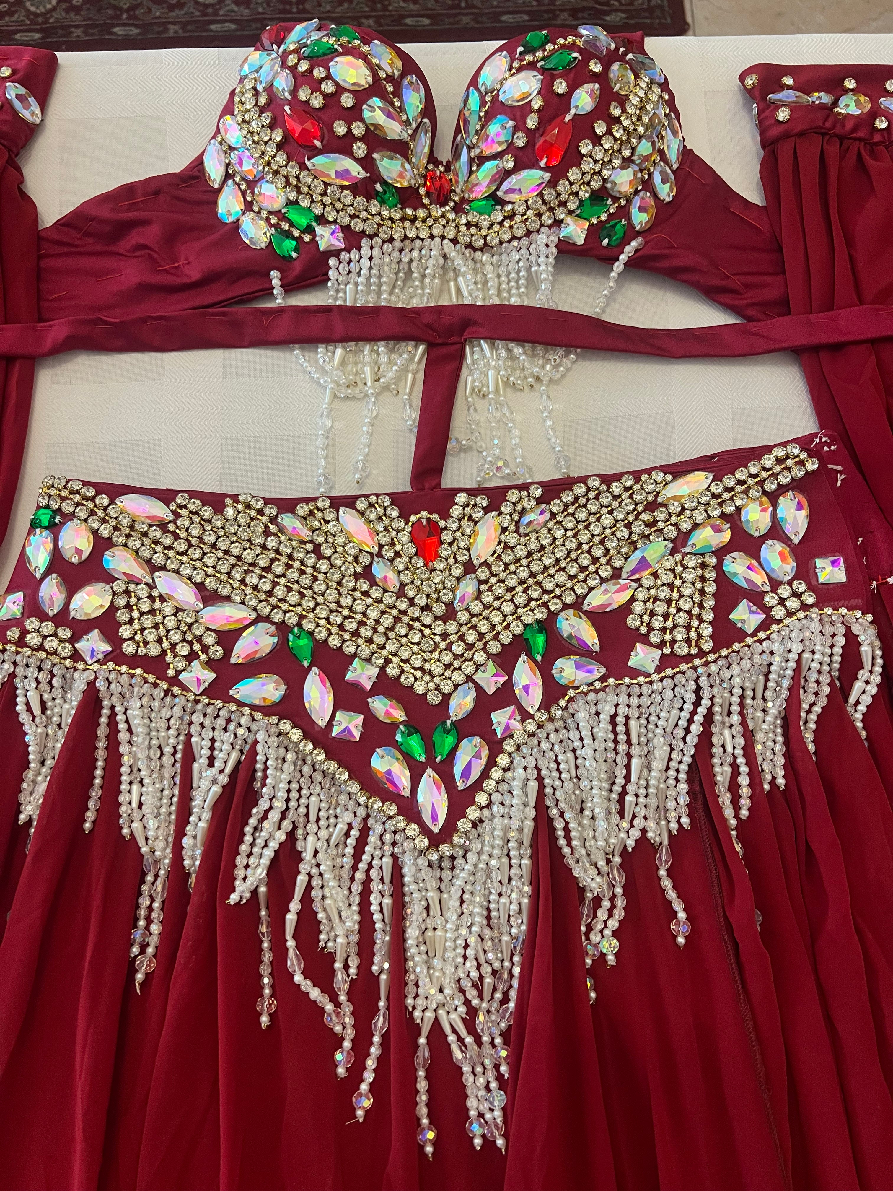 A close-up of a red belly dance costume set featuring a bra top and hip scarf with extensive gold rhinestone embellishments, iridescent teardrop gems, and dangling white pearl bead fringes.