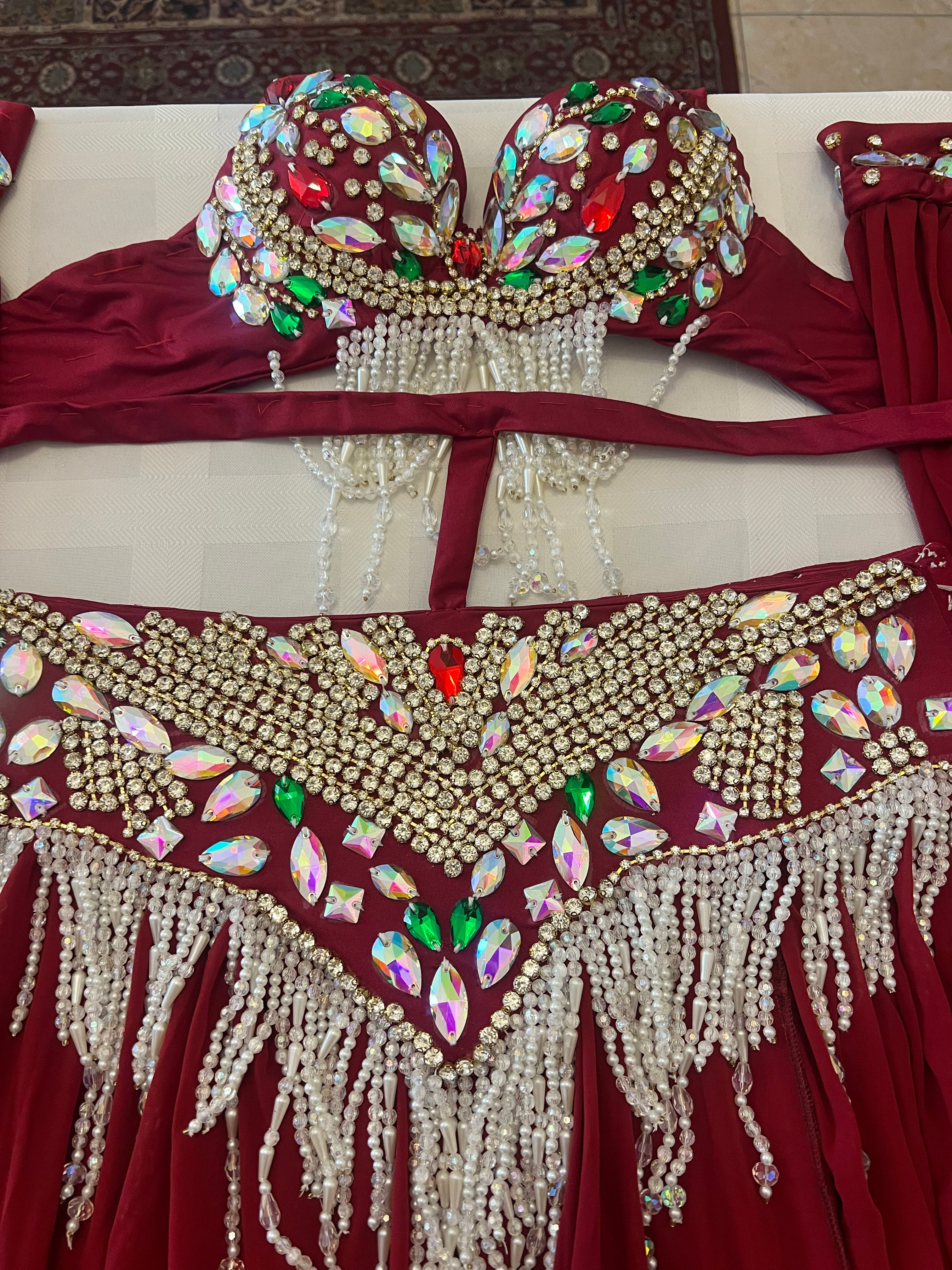A close-up of a belly dance costume set featuring a deep burgundy red fabric with extensive gold rhinestone embellishments, iridescent jewel rhinestones, and hanging white faux pearl bead fringes.