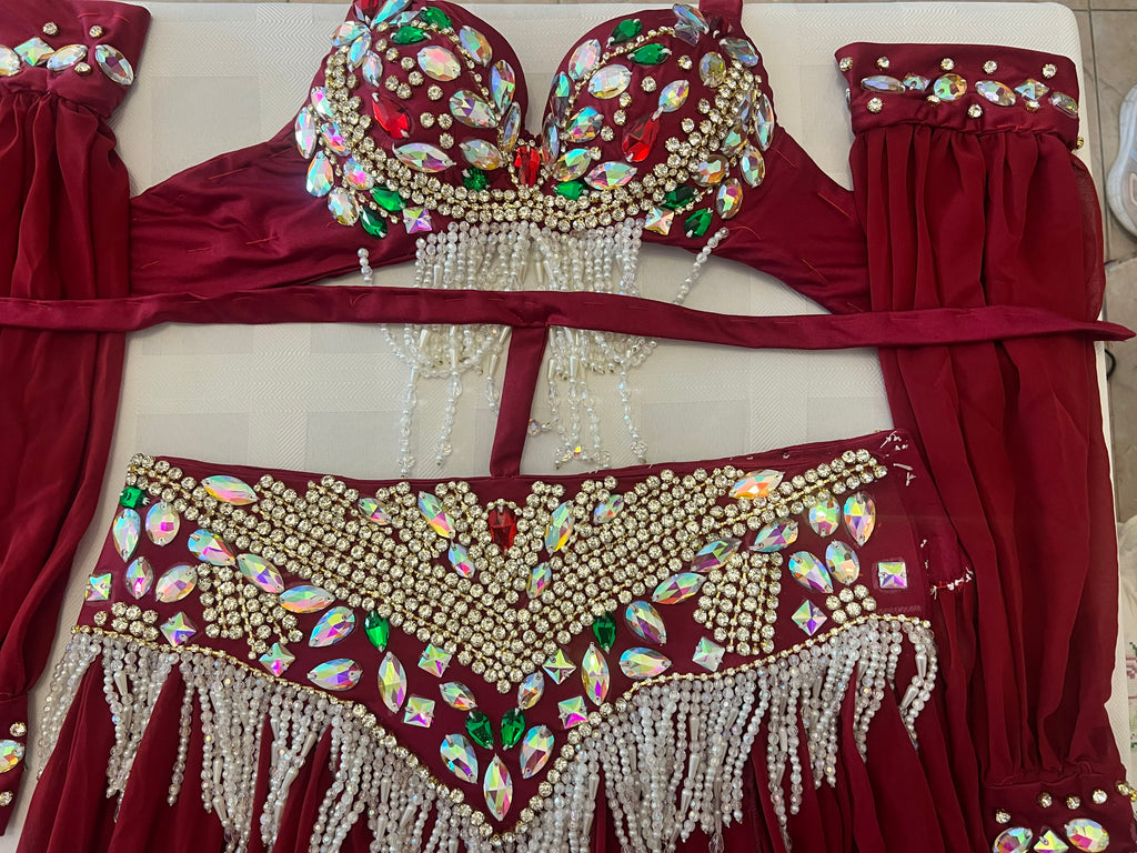 A red belly dance costume set laid flat on a light beige surface, featuring an embellished bra top with rhinestones and bead fringe, a matching waistband with fringe, and coordinating red fabric panels.