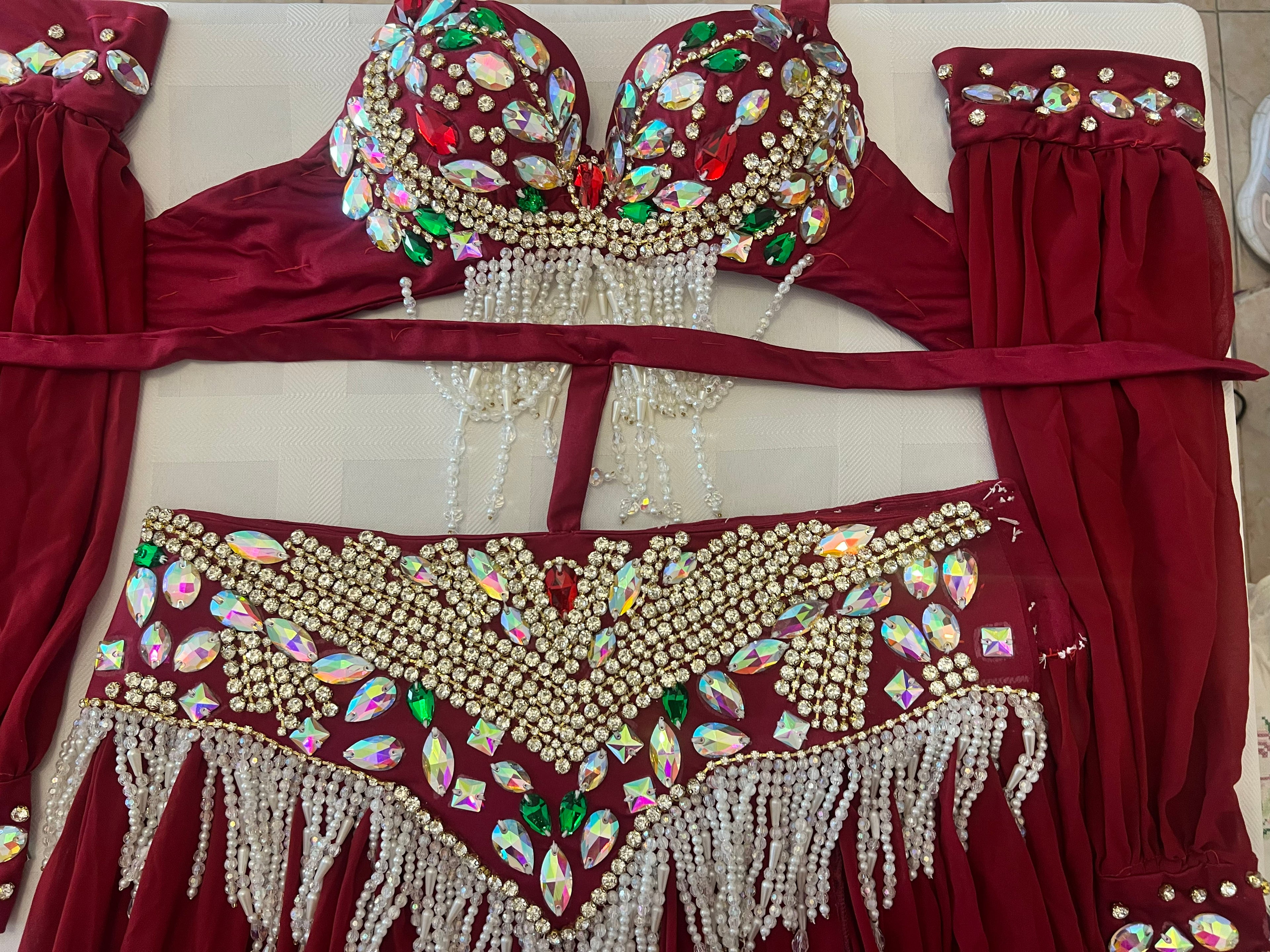 A red belly dance costume set laid flat on a light beige surface, featuring an embellished bra top with rhinestones and bead fringe, a matching waistband with fringe, and coordinating red fabric panels.