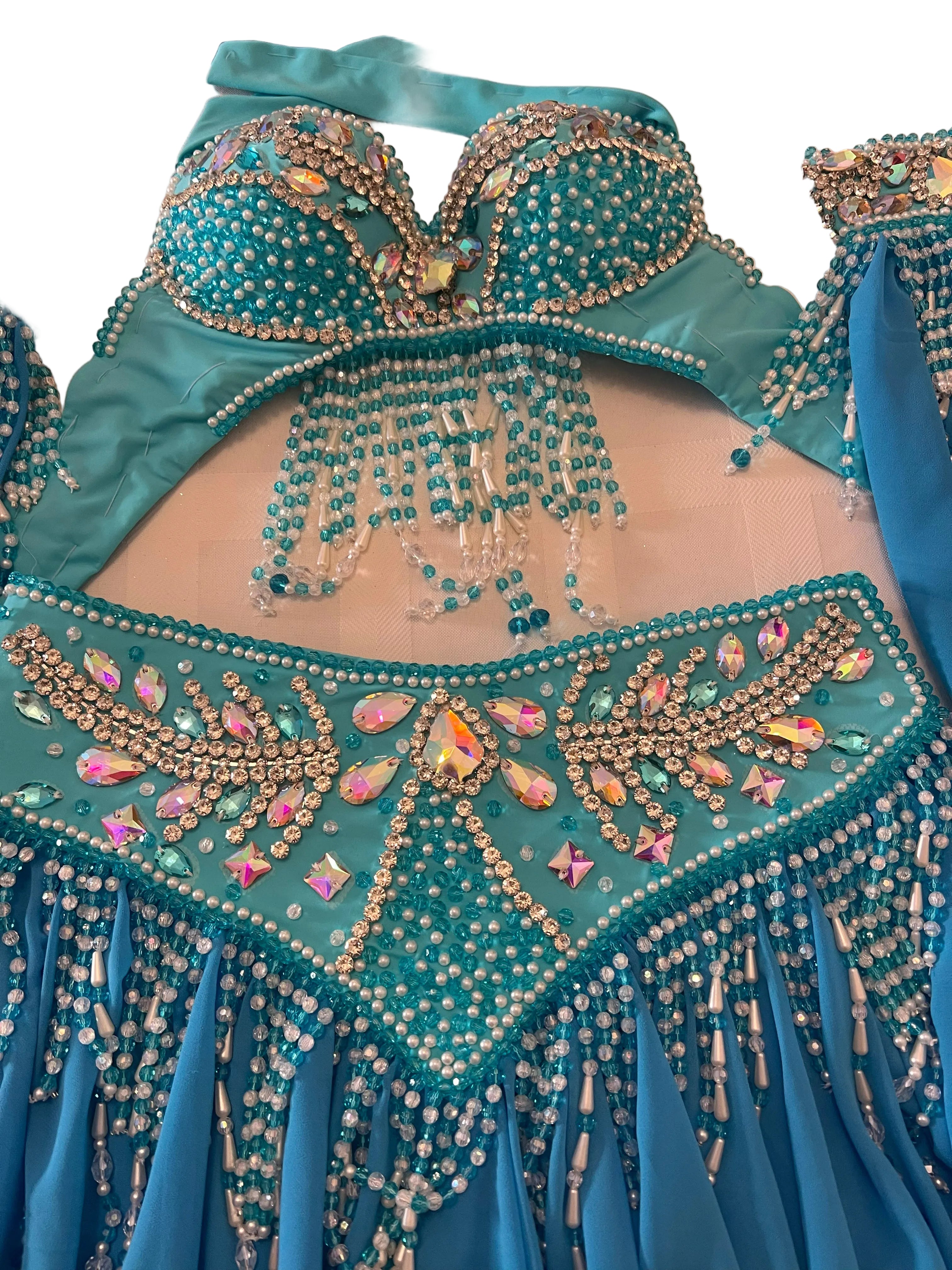 A turquoise and blue belly dance costume set with intricate beadwork, rhinestones, and fringe details, laid out on a light-colored surface.