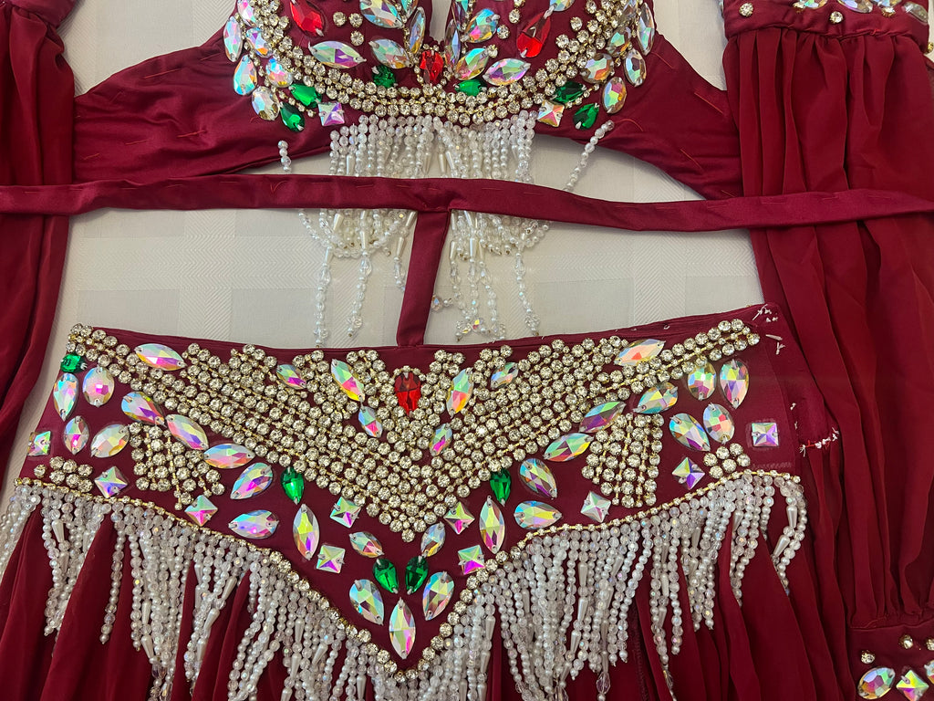 A close-up of a deep red belly dance costume set featuring a bra top and matching hip scarf, both heavily embellished with gold rhinestones, colorful faux gems, and white bead fringe.