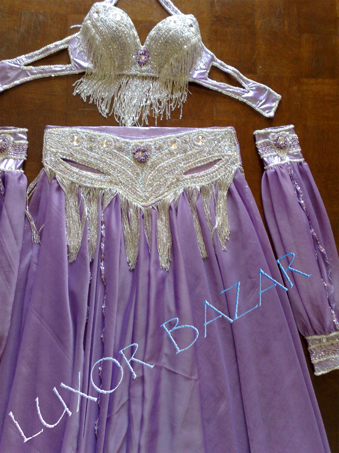 A professional Egyptian belly dance costume with a purple skirt, white chiffon top, and silver decorations including beads and sequins. The costume is displayed on a hanger with the text 'LUXOR BAZAR' visible.