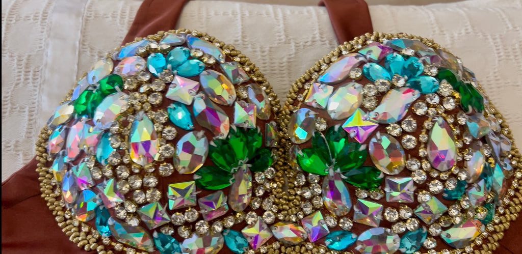 A close-up of a bra cup heavily embellished with large iridescent rhinestones in blue, green, pink, and gold, set against a cream textured fabric background.
