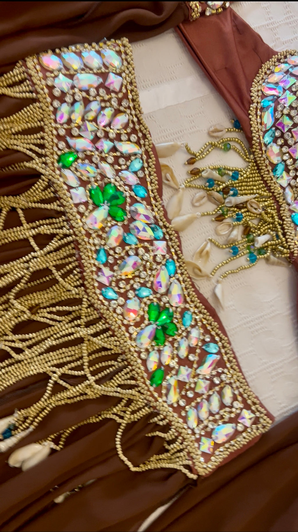 Close-up of a brown embellished garment or accessory featuring a wide decorative panel with gold bead fringe, iridescent rhinestone-like stones, and bright green floral accents on a cream fabric background.