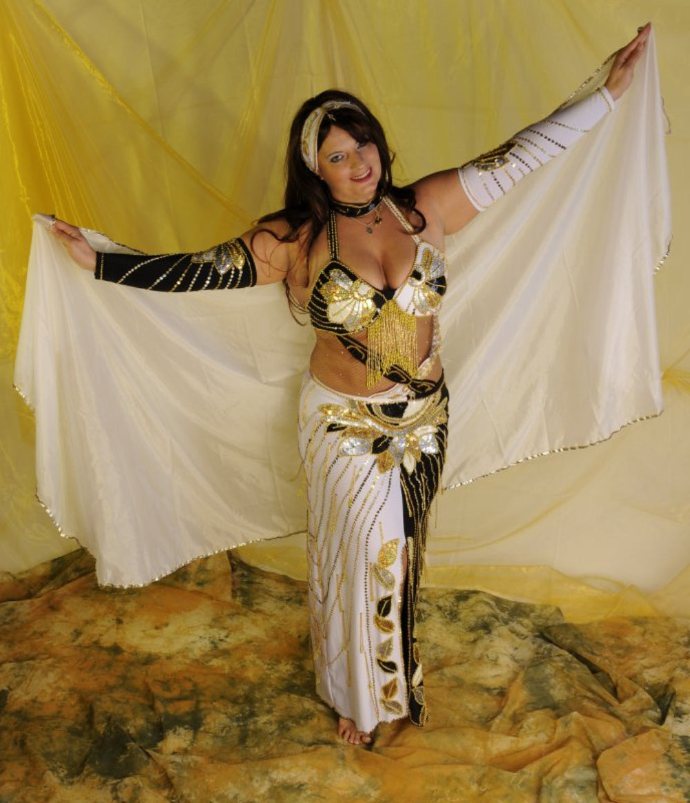 A woman posing in a white and gold belly dance costume with a headband, sleeves, and a skirt, against a yellow background.