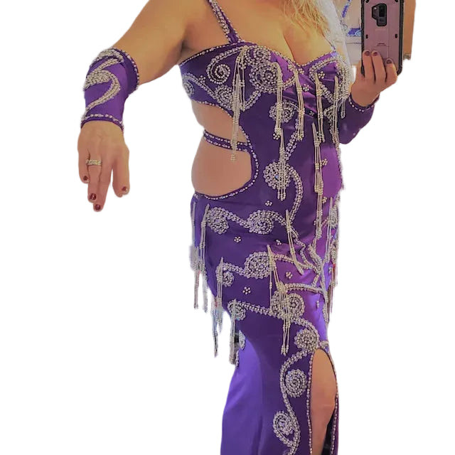 A woman wearing a custom-made purple belly dance costume with sleeves and a headband, posing in a room with a mirror.