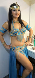 A woman modeling a professional belly dance costume with a blue skirt, gold bra, and accessories, standing in a room with lighting for theatrical or photographic use.