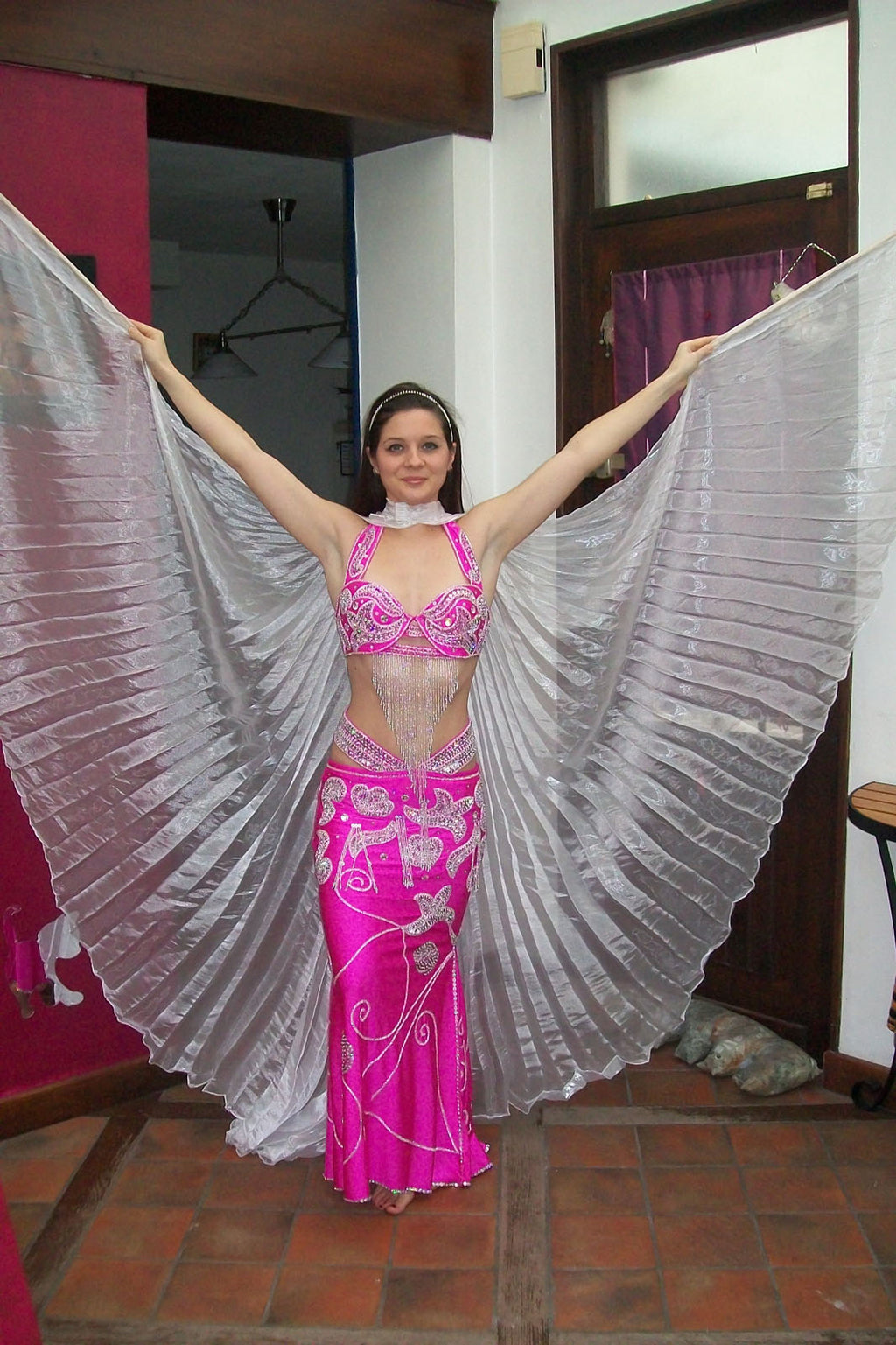 A woman in a professional belly dance costume with a wide skirt, posing with one arm extended upwards. The costume is primarily pink with silver embroidery and a headband.